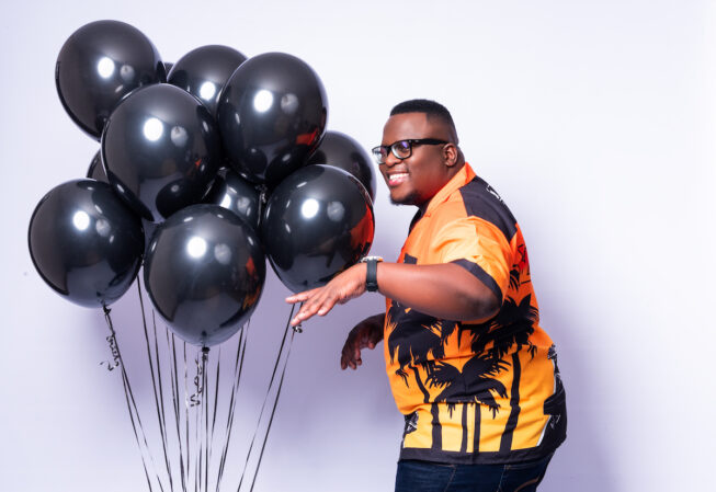 Sol Phenduka in an orange shirt with a bunch of black balloons, getting ready to host the reunion of This Body Works for Me on Showmax