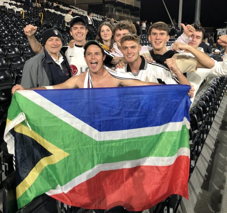 Chairman of the Fulham Supporters Club in SA, James Preston, holding up an SA flag at Fulham's stadium, Craven Cottage along with other fans.