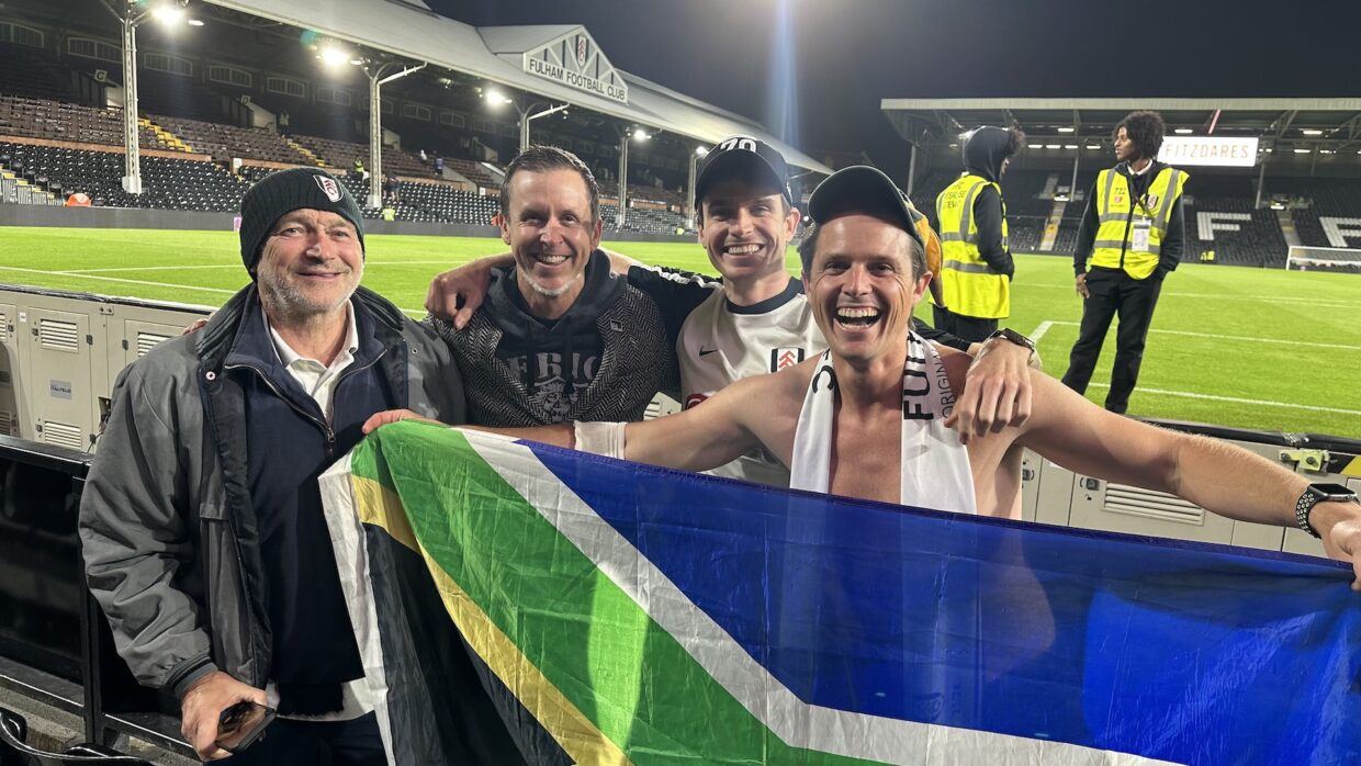 Chairman of the Fulham Supporters Club in SA, James Preston, holding up an SA flag at Fulham's stadium, Craven Cottage along with other fans.